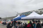 Journalists, videographers, and other interested people under tents record a train passing by on the bridge above the Interstate 5 Freeway & Empire Avenue Interchange